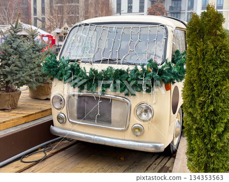 Vintage van decorated with Christmas garland and lights at winter market. Festive urban installation, seasonal decor and holiday atmosphere Vintage van decorated with Christmas garland and lights at winter market. Festive urban installation, seasonal decor and holiday atmosphere 134353563