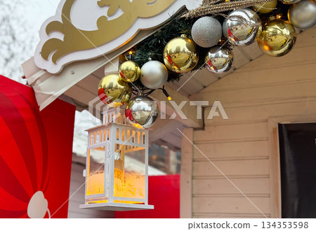 White lantern with warm light hanging under festive roof decor. Christmas market detail, cozy illumination and winter holiday atmosphere White lantern with warm light hanging under festive roof decor. Christmas market detail, cozy illumination and winter holiday atmosphere 134353598