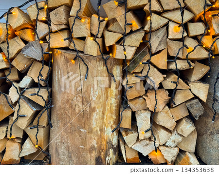 Stacked firewood decorated with warm string lights for Christmas season. Rustic winter background, cozy holiday atmosphere and traditional outdoor styling. 134353638
