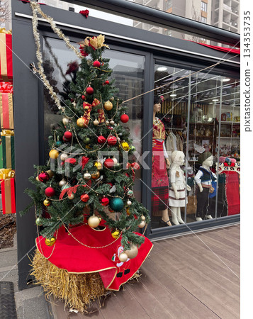 Decorated Christmas tree with red and gold ornaments placed near festive storefront window. Seasonal street decoration, winter celebration, holiday atmosphere and traditional urban display. 134353735