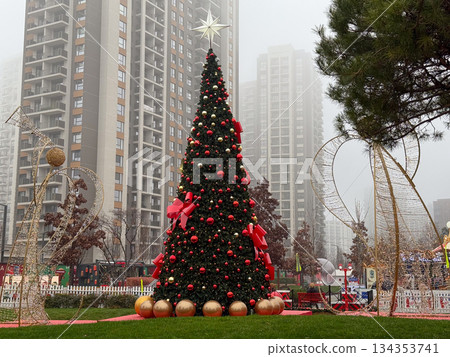 Large Christmas tree with red ornaments and star in modern city square. Winter holiday landmark, seasonal decoration, festive public space and urban celebration. 134353741