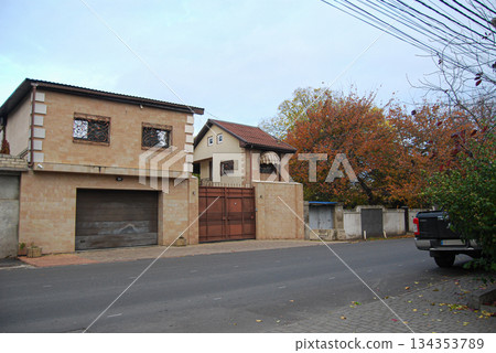 Residential street view with two houses and autumn trees. Two houses on a residential street, featuring light brown facades and patterned windows. 134353789