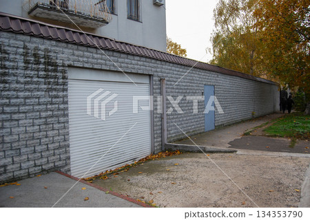 White roll-up garage door in a long grey brick wall with autumn leaves. White roll-up garage door in grey brick wall. The wall extends along a narrow path surrounded by autumn leaves.  134353790