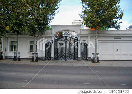 Ornate black gate of a grand white residence. Ornate black wrought iron gate stands as the centerpiece of a grand white residence, featuring intricate scrollwork and decorative patterns. 134353793