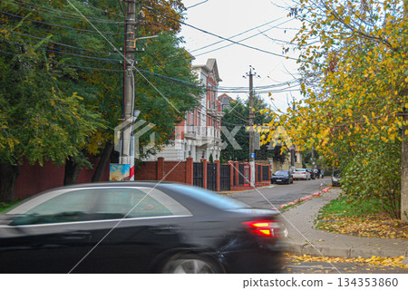 Autumn street scene with a blurry car and ornate building. Street scene in autumn with a motion-blurred black car in the foreground. The street is lined with trees displaying yellow and green leaves. Autumn street scene with a blurry car and ornate building. Street scene in autumn with a motion-blurred black car in the foreground. The street is lined with trees displaying yellow and green leaves. 134353860