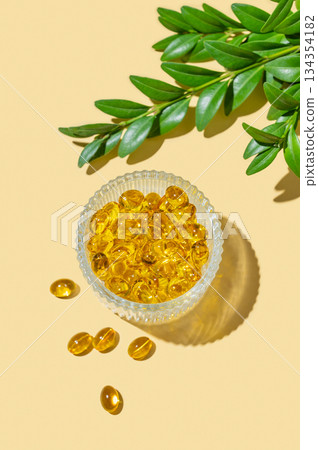 Yellow softgel vitamins capsules in a bowl on a bright background with green leaves and shadow. Yellow softgel vitamins capsules in a bowl on a bright background with green leaves and shadow. 134354182