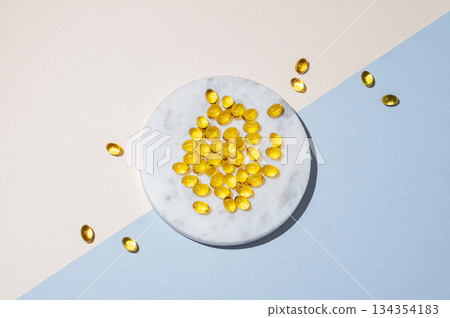 Yellow softgel vitamins capsules on a white marble plate on a blue background with shadow. Yellow softgel vitamins capsules on a white marble plate on a blue background with shadow. 134354183