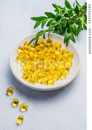 Yellow softgel vitamins capsules on a white plate with green leaves on a blue background Yellow softgel vitamins capsules on a white plate with green leaves on a blue background 134354184