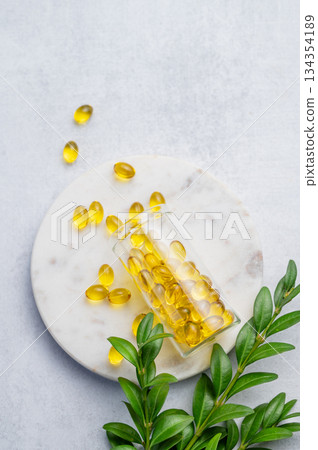 Yellow softgel vitamins capsules in a bottle on a marble standon a light background with green leaf. Yellow softgel vitamins capsules in a bottle on a marble standon a light background with green leaf. 134354189