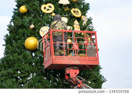 Worker decorates a tall city Christmas tree with golden ornaments using a lift, evergreen branches against a winter sky in Moscow 134354708