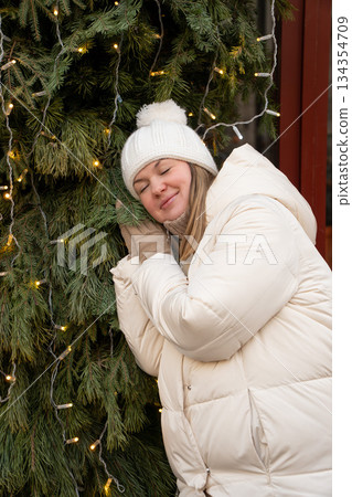 Smiling woman in a white winter jacket and knitted hat leans against a decorated evergreen with warm fairy lights, creating a calm, cozy holiday mood Smiling woman in a white winter jacket and knitted hat leans against a decorated evergreen with warm fairy lights, creating a calm, cozy holiday mood 134354709