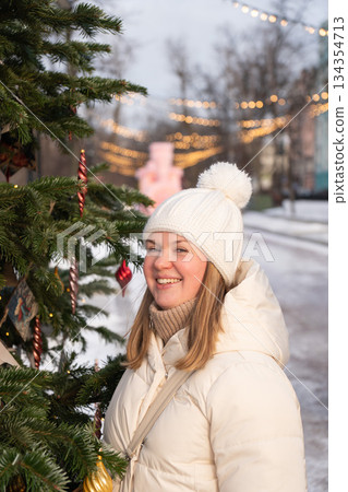 Smiling young woman in a white winter jacket and knitted hat stands outdoors near a Christmas tree with warm city lights in the background Smiling young woman in a white winter jacket and knitted hat stands outdoors near a Christmas tree with warm city lights in the background 134354713