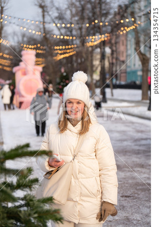 A smiling woman in a white winter coat and knitted hat walks along a festive city boulevard with string lights and snow, holding a smartphone 134354715