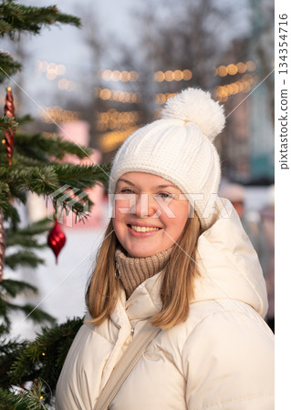 Smiling young woman in a white winter jacket and knitted hat stands outdoors near a Christmas tree with warm city lights in the background 134354716