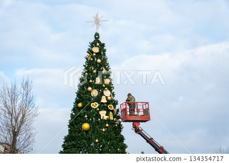Worker decorates a tall city Christmas tree with golden ornaments using a lift, evergreen branches against a winter sky in Moscow Worker decorates a tall city Christmas tree with golden ornaments using a lift, evergreen branches against a winter sky in Moscow 134354717