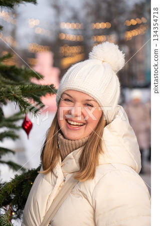 Smiling young woman in a white winter coat and knitted hat stands by a decorated Christmas tree with warm lights on Tverskoy Boulevard in Moscow 134354718