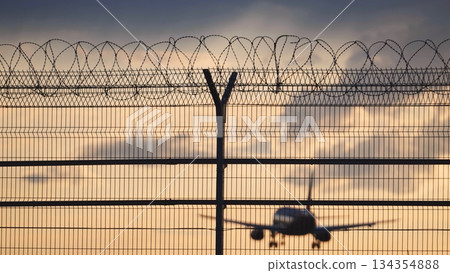 Airplane landing behind airport fence with barbed wire at sunset Airplane landing behind airport fence with barbed wire at sunset 134354888
