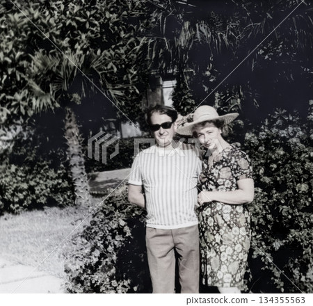 Vintage portrait of a happy married couple in a summer garden. The woman wears a straw hat. The man wears dark glasses. Retro photo, 1971. 134355563