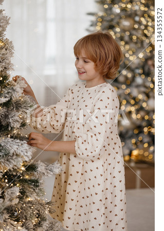 Side view of Caucasian blonde girl decorating Christmas tree in cozy room Side view of Caucasian blonde girl decorating Christmas tree in cozy room 134355572