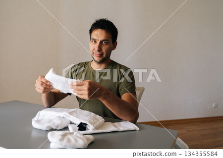 Maintenance worker carefully checking microfiber cloths, meticulously preparing cleaning supplies for professional sanitation workflow sitting at table at looking at camera. Concept of household. Maintenance worker carefully checking microfiber cloths, meticulously preparing cleaning supplies for professional sanitation workflow sitting at table at looking at camera. Concept of household. 134355584