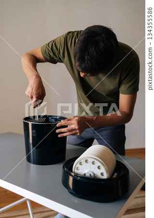 Vertical shot of technician disassembling and performing maintenance on modern black air humidifier using screwdriver, showcasing internal components and filter on table. Concept of indoors hygiene. Vertical shot of technician disassembling and performing maintenance on modern black air humidifier using screwdriver, showcasing internal components and filter on table. Concept of indoors hygiene. 134355586