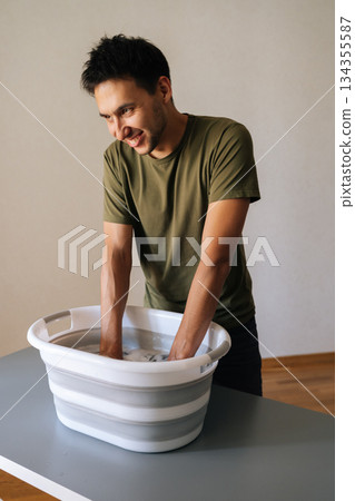 Vertical portrait of positive young man washing clothes by hand in plastic basin, wringing out water performing domestic task, smiling looking away. Concept of housework, housekeeping and household. Vertical portrait of positive young man washing clothes by hand in plastic basin, wringing out water performing domestic task, smiling looking away. Concept of housework, housekeeping and household. 134355587