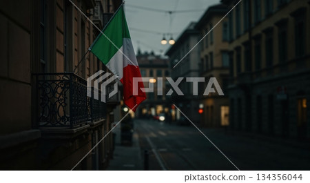 An Italian flag with vibrant green, white, and red stripes hangs from an ornate dark metal balcony on a historic building facade above a blurred European city street at dusk. 134356044