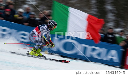 A male alpine skier in a grey and yellow suit carves down a snowy slope, kicking up powder with an Italian flag and cheering spectators blurred in the background. A male alpine skier in a grey and yellow suit carves down a snowy slope, kicking up powder with an Italian flag and cheering spectators blurred in the background. 134356045