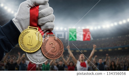 A gloved hand proudly displays gold, silver, and bronze medals with red ribbons against a blurred stadium background featuring a cheering crowd and the Italian flag. 134356056