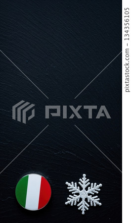 A round Italian flag badge and a sparkling white snowflake ornament are neatly arranged on a dark, textured slate background, providing ample copy space. 134356105