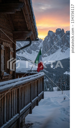 A rustic wooden chalet balcony covered in fresh snow with an Italian flag fluttering, set against a dramatic alpine mountain range and a vibrant sunset sky. 134356117