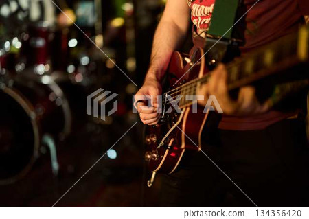 Guitarist Plays Energetically, Warm Amber Lights Highlight Guitarist Strumming Over Drums In Background Guitarist Plays Energetically, Warm Amber Lights Highlight Guitarist Strumming Over Drums In Background 134356420