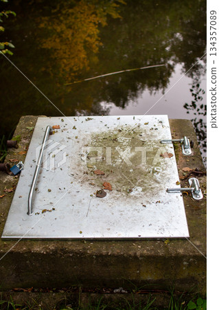 Metal cover rests on a stone platform near a calm water surface surrounded  134357089