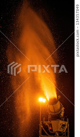 A powerful snow cannon sprays glowing orange particles into the dark night sky, illuminated brightly by a warm artificial light source. 134357849