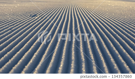 Perfectly groomed white snow on a pristine ski slope at sunrise, featuring crisp parallel corduroy patterns illuminated by warm golden light and casting long shadows. 134357860