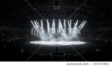 An empty large arena stage is dramatically lit by multiple bright spotlights cutting through atmospheric smoke, creating a powerful, expectant mood. 134357861