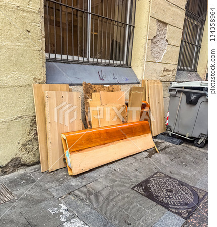 Stack of discarded wooden boards and furniture near building wall and garbage bin. Urban waste, recycling, and renovation concept in city street.. High quality photo 134358964