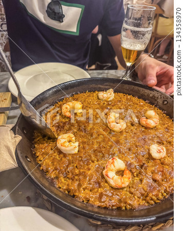 Close-up of Spanish paella with shrimp and metal spoon on table beside glass of beer. Authentic Mediterranean cuisine, dining, and travel concept. High quality photo 134358970