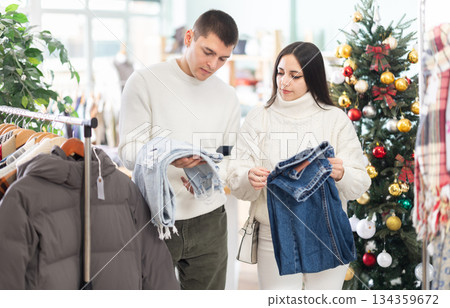 Married couple choose jeans against background of Christmas tree in clothing store Married couple choose jeans against background of Christmas tree in clothing store 134359672