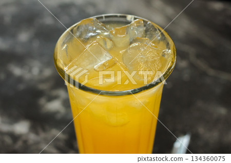 Macro shot of bright orange tropical refreshment drink with melting ice cubes in a tall glass, set against a blurred dark background. 134360075