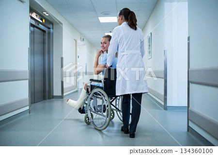 Doctor assisting patient in wheelchair in hospital hallway. A hospital hallway with bright lighting and smooth flooring. Doctor assisting patient in wheelchair in hospital hallway. A hospital hallway with bright lighting and smooth flooring. 134360100