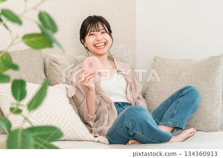 一位年輕的亞裔女性坐在客廳的沙發上吃甜甜圈。 134360913