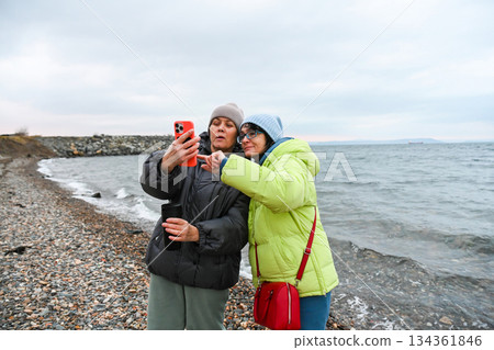 Two Russian friends aged 50 and 60 review photos during a spring walk by the sea, sharing guidance, support and mindful learning in a calm moment. 134361846