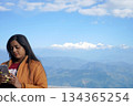 Indian woman in winter jacket using phone against Himalayan mountain range background 134365254