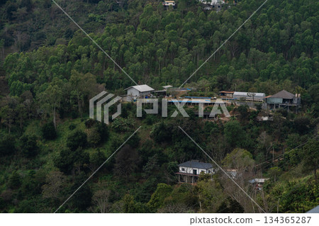 Tranquil hillside village surrounded by lush green forest featuring residential buildings and remote cabins in a peaceful natural setting 134365287