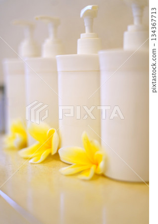 Soft focus image featuring white pump bottles arranged in a row with fresh yellow plumeria flowers at the base. Clean, spa-like scene ideal for beauty, skincare, and wellness branding. Soft focus image featuring white pump bottles arranged in a row with fresh yellow plumeria flowers at the base. Clean, spa-like scene ideal for beauty, skincare, and wellness branding. 134367713