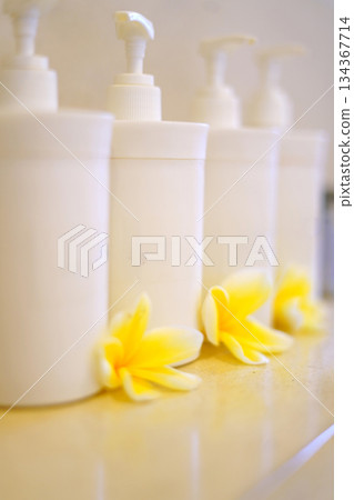 Soft focus image featuring white pump bottles arranged in a row with fresh yellow plumeria flowers at the base. Clean, spa-like scene ideal for beauty, skincare, and wellness branding. 134367714