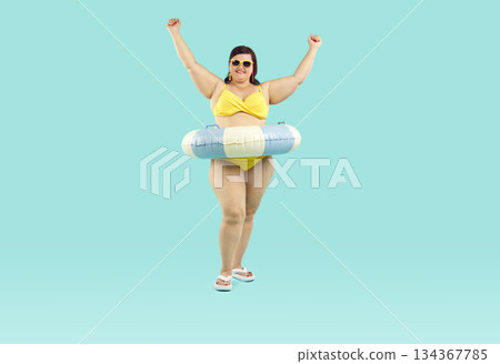 Funny fat woman in sunglasses in rubber ring with hands up on studio blue background. 134367785