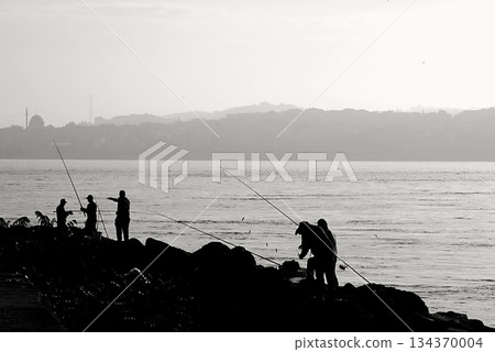 Silhouettes of Bosphorus fishermen in the early morning. A black-and-white, high-contrast image that captures the serene solitude of recreational fishing in Istanbul. Silhouettes of Bosphorus fishermen in the early morning. A black-and-white, high-contrast image that captures the serene solitude of recreational fishing in Istanbul. 134370004