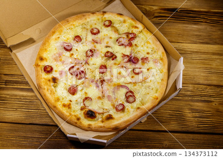 Delicious fresh pizza in cardboard box on a wooden table. Top view. Concept for home delivery of food, fast food, delivery of pizza Delicious fresh pizza in cardboard box on a wooden table. Top view. Concept for home delivery of food, fast food, delivery of pizza 134373112